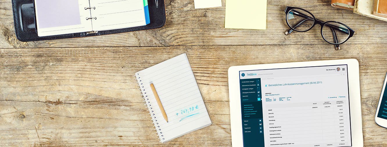 Tablet auf einem Schreibtisch mit TASSlink: Betriebliches Lohnkostenmanagement für einen Mitarbeiter, Bausteine wie Warengutschein, Verpflegungszuschuss, Kindergartenzuschuss, Fahrtkostenzuschuss, Gesundheitsförderung und E-Bike mit Beträgen, daneben eine Notiz mit 264,98 Euro.